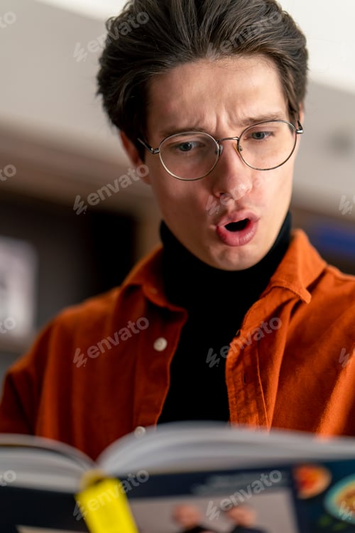 Preview: young guy in glasses stands near the bookshelves holds reads a book enthusiastically marvels story