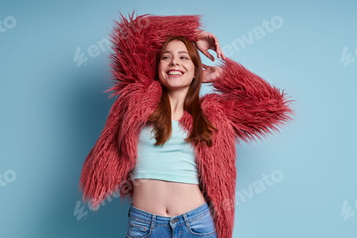 Preview: Beautiful young woman in fluffy coat smiling while standing against blue background