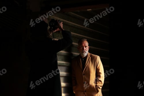 Preview: Photographer in action, making a photo of a man with a grey beard, in a shadowy place.