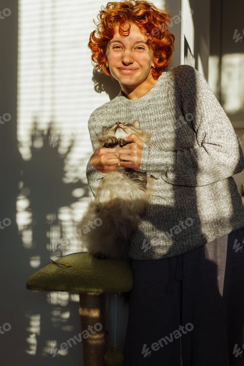 Preview: portrait red-haired curly young woman with pet gray fluffy cat in arms, lit by bright sun