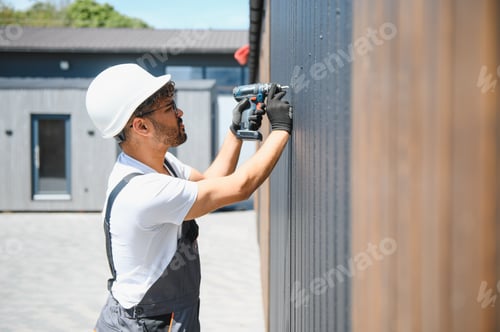 Preview: Construction worker using cordless drill installing siding on prefabricated building