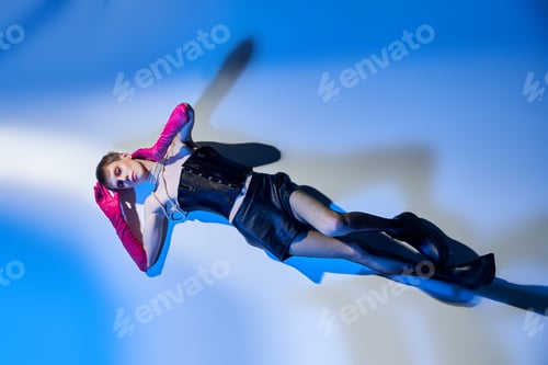 Preview: top view, queer model in corset and pink gloves lying on blue backdrop, lgbtq, bold style, fashion