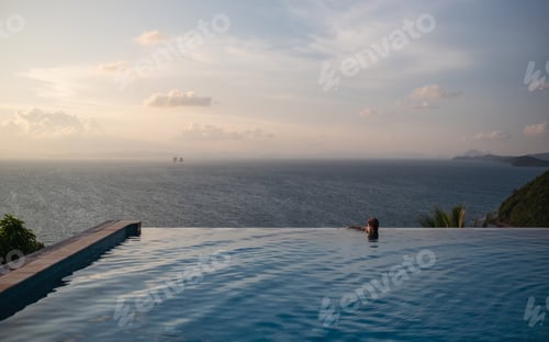 Preview: A young woman relaxing in infinity swimming pool and looking at a beautiful sunset and the sea view