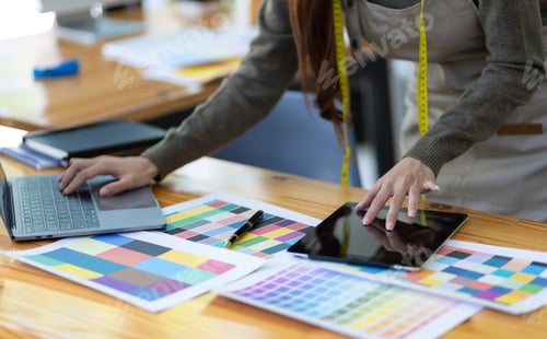 Preview: Fashion designer in design studio working with laptop and fabric color samples on desk.