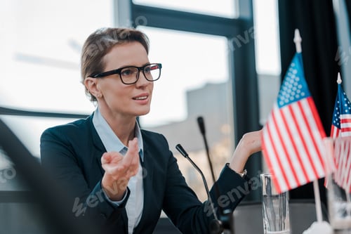 Preview: beautiful diplomat in eyeglasses gesturing while talking near microphone and american flags