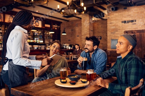 Preview: Group of young people choosing order from digital menu while talking to a waitress in a pub.