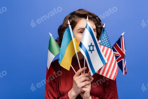 Preview: young woman looking at camera behind various international flags isolated on blue
