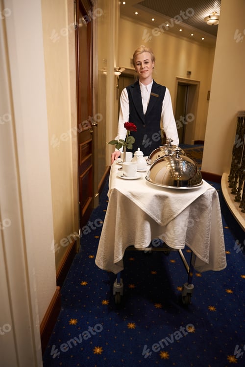 Preview: Young waitress on trolley carries hot food along hotel corridor