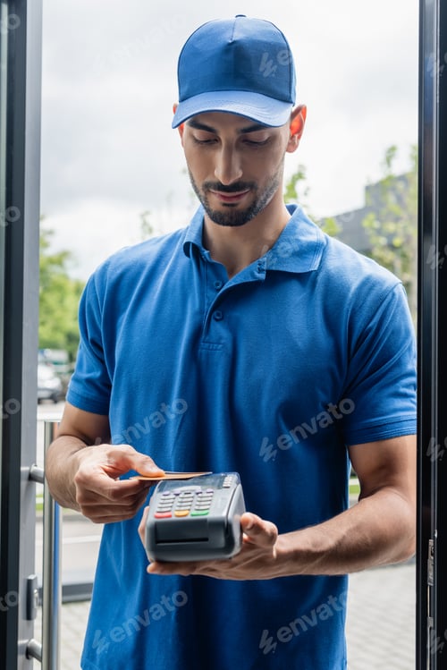 Preview: Arabian courier holding payment terminal and credit card near door