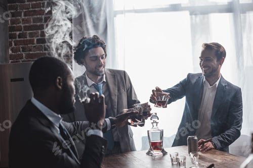 Preview: three smiling multiethnic businessmen smoking cigars and clinking glasses of whiskey