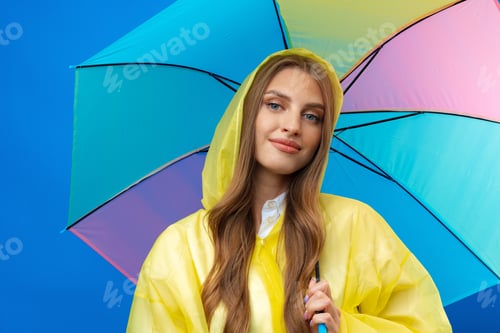 Preview: Young woman in yellow raincoat with rainbow umbrella against blue background in studio