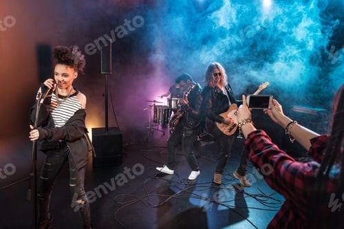 Preview: Young woman photographing rock and roll band performing hard rock music on stage