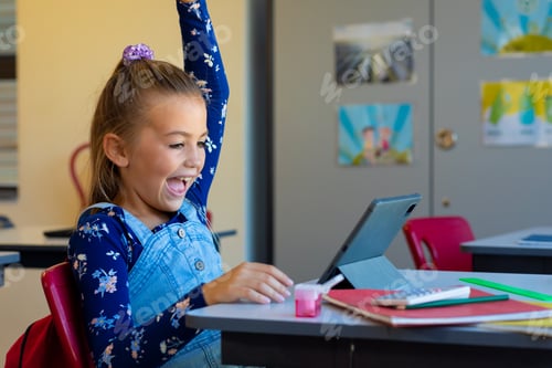 Preview: Happy caucasian girl raising her hand while having videocall on laptop in class at elementary school