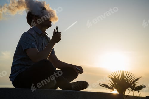 Preview: vaping young man with, produces vapor on sunset sky background at the sea coast promenade, place for