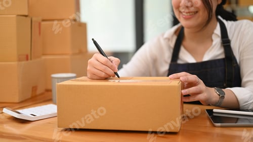 Preview: Female small business entrepreneur writing address on cardboard box.