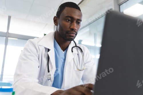 Preview: African american male doctor wearing lab coat and stethoscope, using laptop at hospital