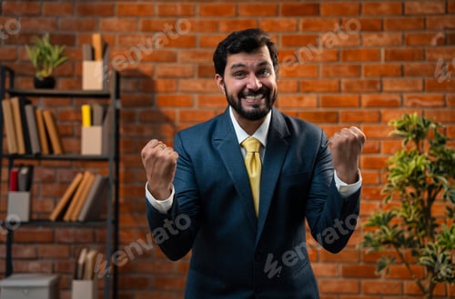 Preview: Smiling Indian businessman gives thumbs up while posing in office with brick wall background