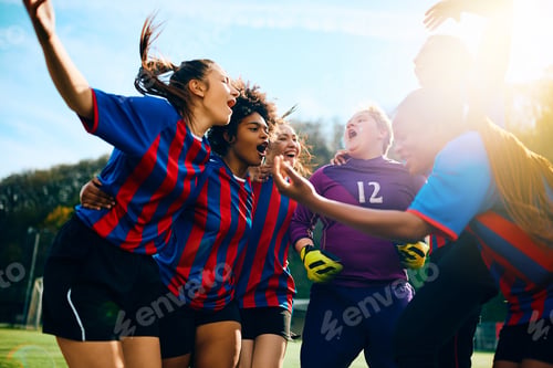 Preview: Cheerful female soccer players shouting while celebrating victory on playing field.