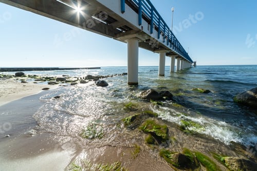 Preview: Pier on coast. Surf waves with sea foam and seaweed on tourist beach. Beautiful coast in sunlight