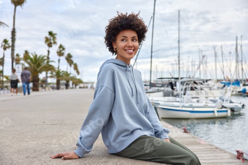 Preview: Horizontal shot of cheerful woman poses at pier alone looks happily away enjoys marine views and goo