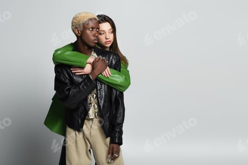 Preview: brunette woman embracing blonde african american man in trendy clothes and looking away on grey