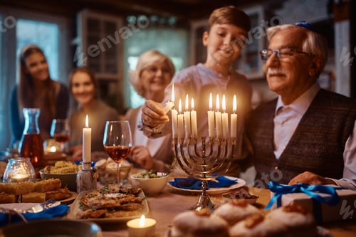 Preview: Close up of Jewish boy lighting menorah while celebrating Hanukkah with his family at dining table.