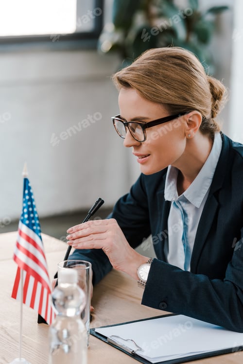 Preview: selective focus of attractive diplomat in eyeglasses touching microphone near clipboard and american