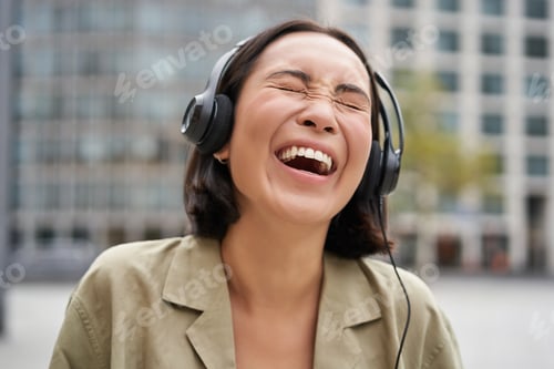 Preview: Portrait of happy asian woman in headphones, enjoys music while walking in city, smiling and