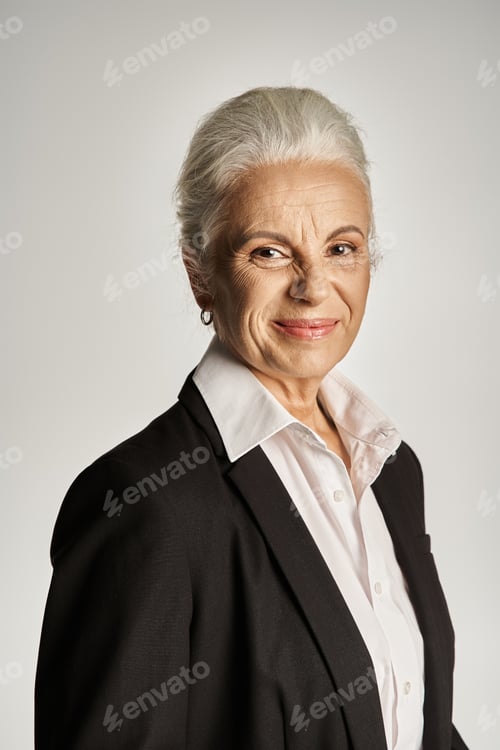 Preview: business portrait of happy middle aged woman with grey hair posing in formal wear on grey backdrop