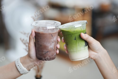Preview: Hands of teenage girls holding glass of ice chocolate and matcha green tea together at cafe.