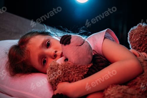 Preview: frightened child embracing teddy bear while lying in bed and looking at camera