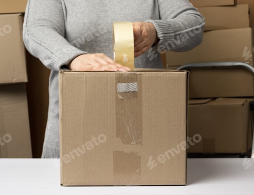 Preview: Woman in a gray sweater holds a roll of duct tape and packs brown cardboard boxes on a white table