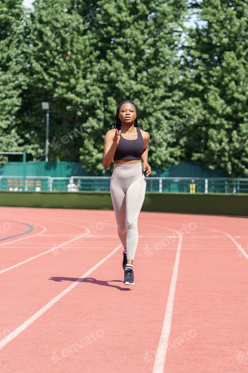 Preview: Focused sportive woman runs on red track of big stadium against city park with lush green trees
