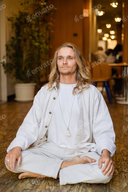 Preview: young and stylish man with long fair hair looking away while meditating in easy pose in cottage