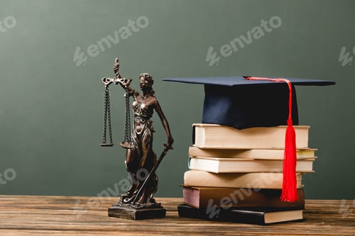 Preview: stack of books, themis statuette and academic cap on wooden surface on grey
