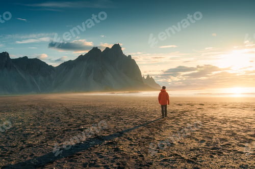 Preview: Sunrise over Vestrahorn mountain with a tourist man walking on black sand beach in summer