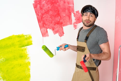 Preview: young man in working overall holding paint rollers and doing shrug gesture in front of painted wall