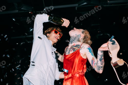 Preview: handsome man in sunglasses pouring champagne from bottle in mouth of girl in red dress on black