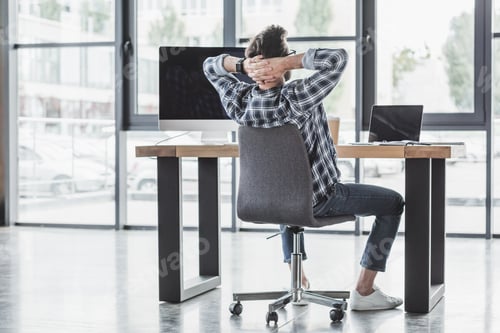 Preview: back view of young programmer sitting with hands behind head at workplace
