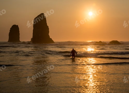 Preview: Surfer walking out of the sea with sun setting behind rock in waves on beach.