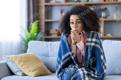 Preview: Young woman feeling cold and wrapped in a blanket while sitting on a cozy couch in living room