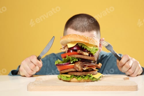 Preview: Young man with cutlery and tasty huge burger at table