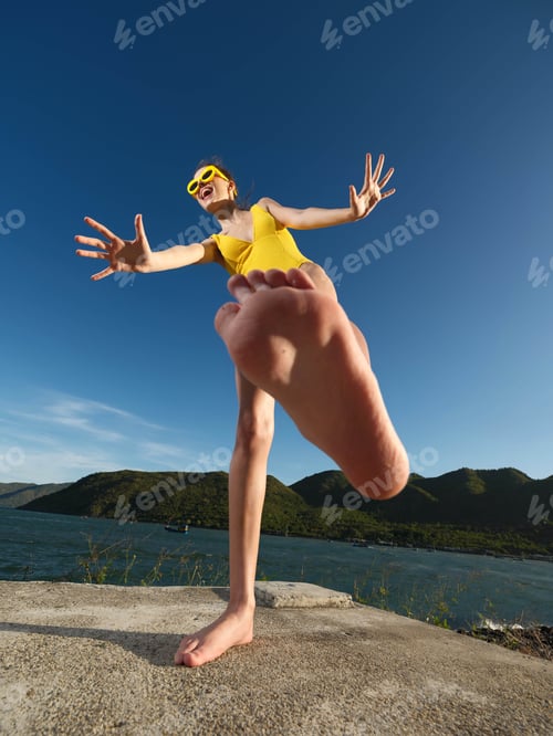 Preview: Young woman in yellow swimsuit, legs raised, near serene water with scenic mountain backdrop