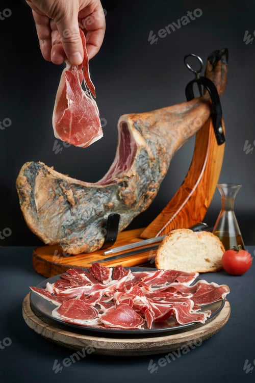 Preview: Chef holding slice of iberian ham from ham leg with sliced ham, bread, tomato and olive oil