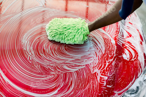 Preview: Closeup cropped image of hand of African man with green sponge washing his red car