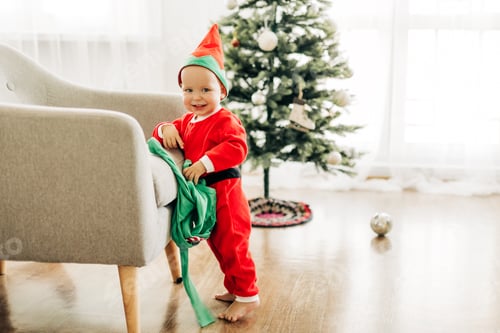 Preview: A little boy kid in a red suit stands near a chair against the background of a decorated Christmas