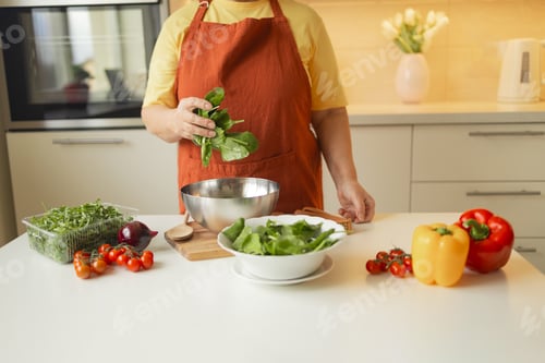 Preview: Senior 60s blond smiling woman holding a knife and fresh spinach whole preparing a bowl of healthy