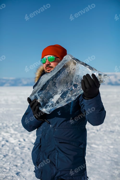Preview: Happy smiling man wearing winter blue jacket and red hat holding piece of transparent crushed ice