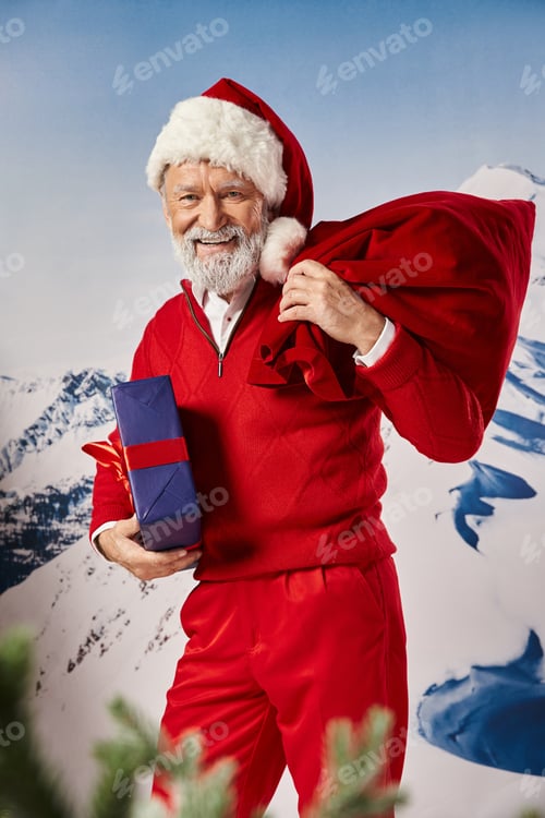 Preview: jolly white bearded man in Santa costume posing with present bag with snowy backdrop, winter concept