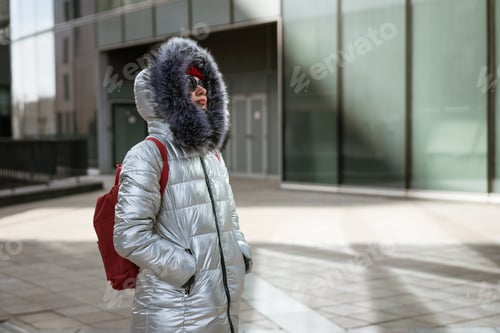 Preview: Happy young woman in winter jacket with backpack near building